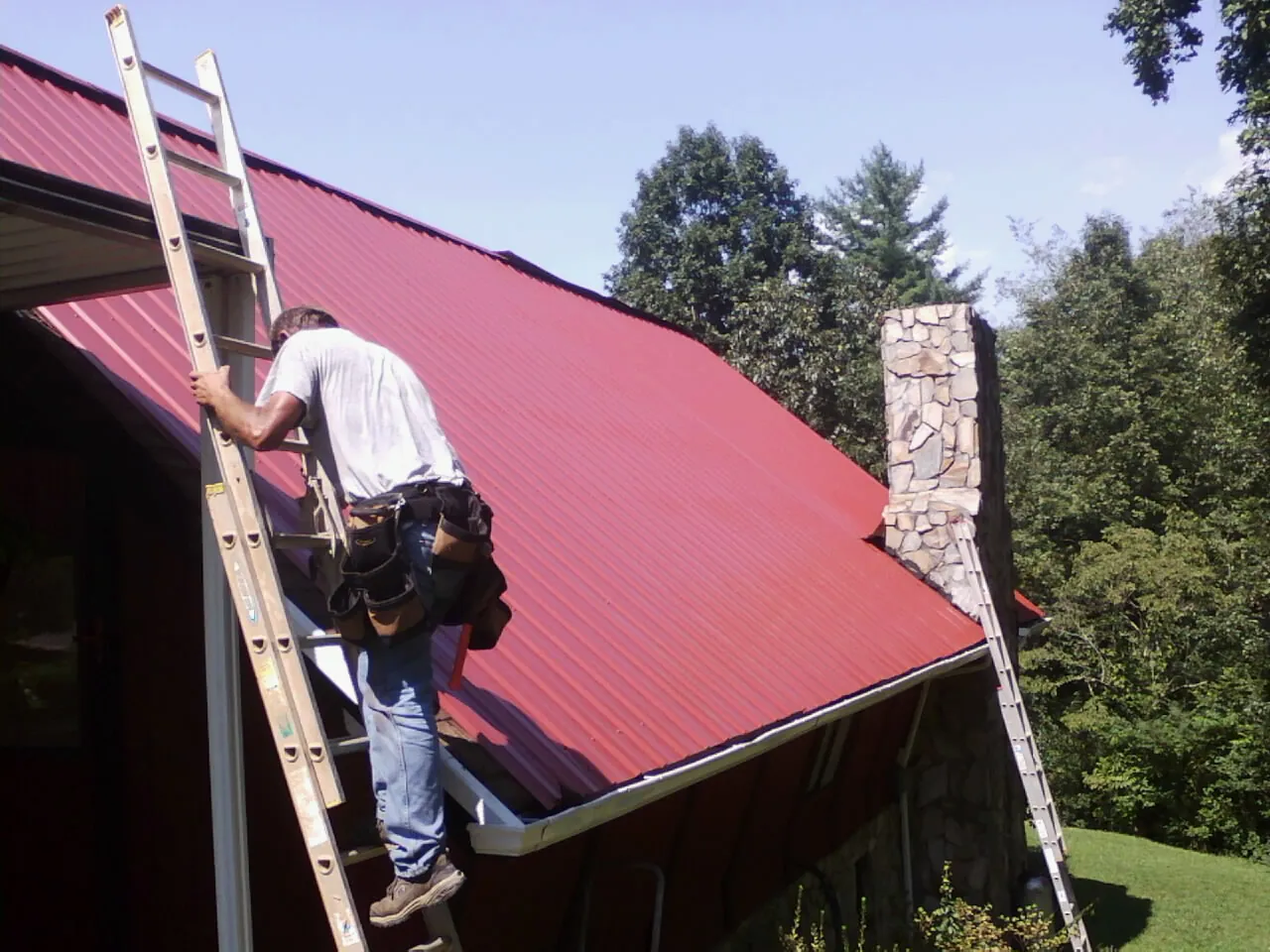 Roofer on a ladder beside a red metal roof and stone chimney — Soffit & Fascia Repair by Roofing Brothers in Leander